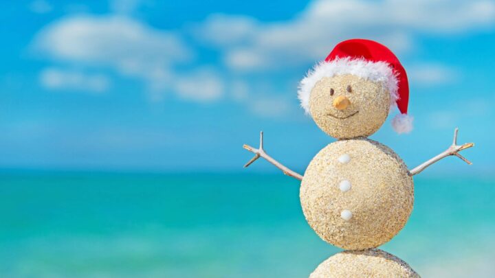snowman made of sand with red Santa hat on against turquoise blue beach water and clouds in sky