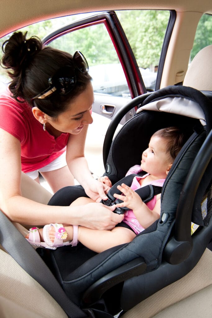 Happy smiling mother placing baby in car seat and closing belt for safety.