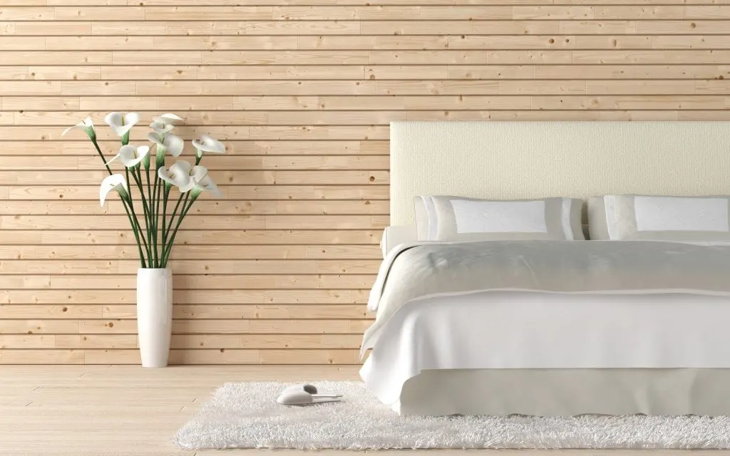 minimalist bedroom with white sheets against a wooden slat wall with a flower vase on the floor