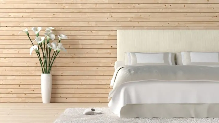 minimalist bedroom with white sheets against a wooden slat wall with a flower vase on the floor