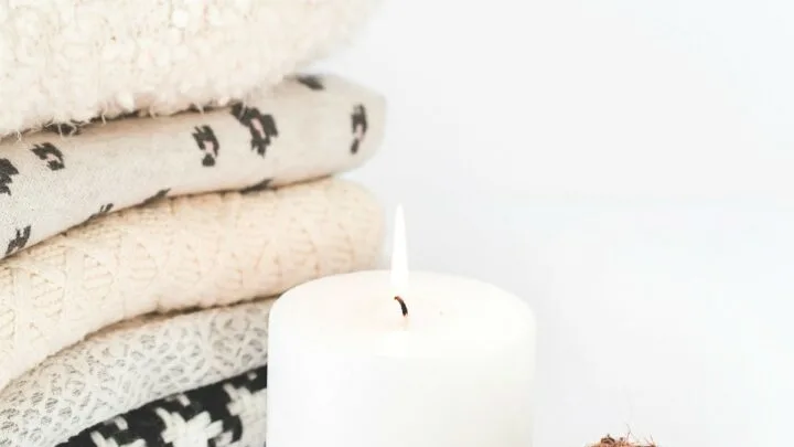 cozy winter sweaters stacked up next to white candle with flame and two pinecones