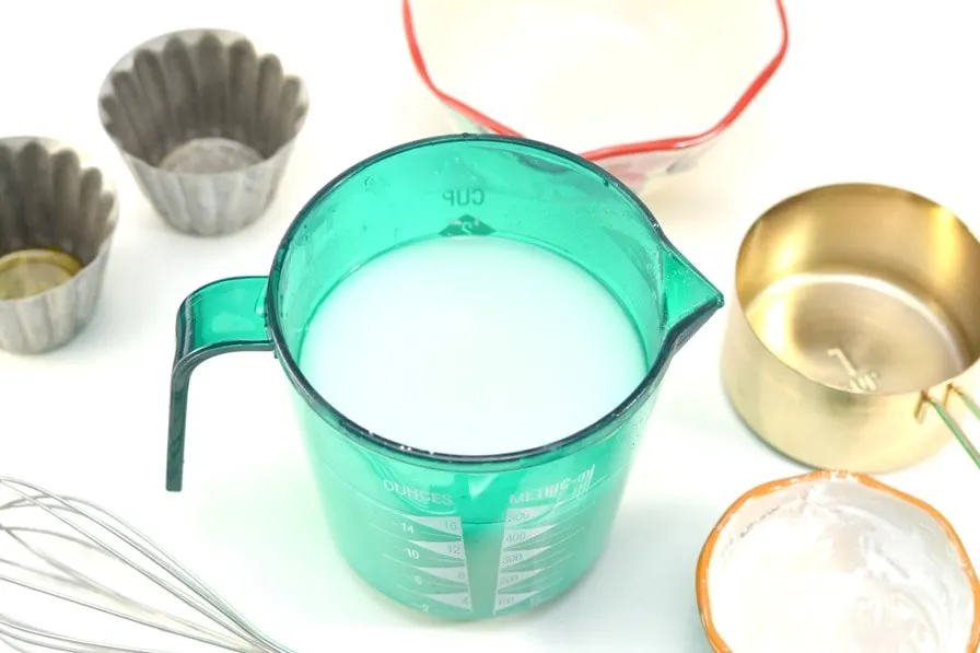 blue measuring cup with cloudy white glass cleaner mixture inside and measuring cups surrounding on white countertop