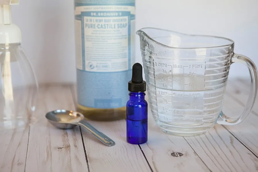 glass measuring cup wth water, stainless steel measuring spoon, blue glass essential oil bottle and castile soap on wooden table