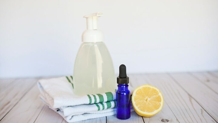 plastic foaming hand soap dispenser on wood table with dishtowel, blue glass essential oilbottle and a slice of lemon