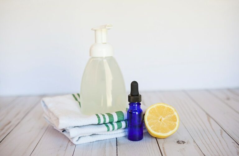plastic foaming hand soap dispenser on wood table with dishtowel, blue glass essential oilbottle and a slice of lemon