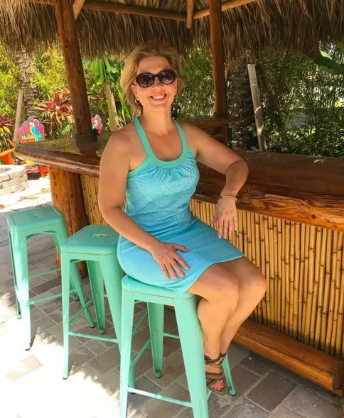 woman sitting down on a teal metal stool wearing teal dress by tiki bar