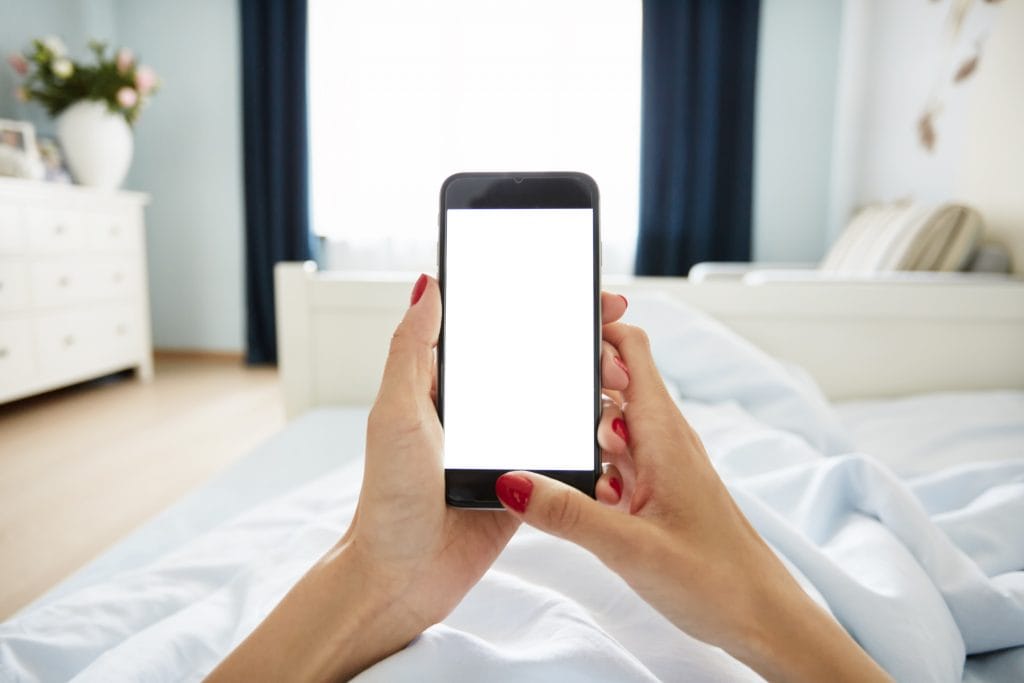 Cropped view of female hands holding smart phone with blank screen for your text. Woman reading messages on cell phone with home interior background while resting in bed.