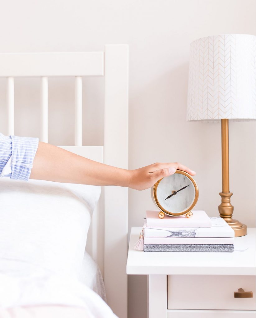 woman with hand on old fashioned bedside clock on night table with lamp against white wall
