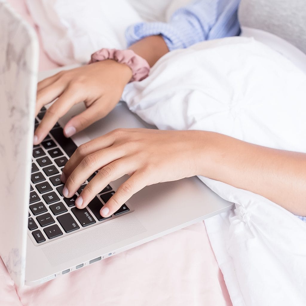 woman typing on laptop computer in bed with white sheets and pink duvet