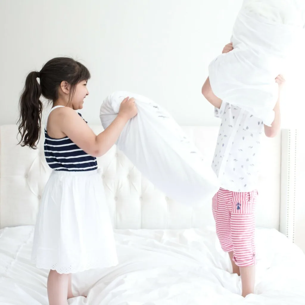 two children jumping on white comforter on bed throwing pillows