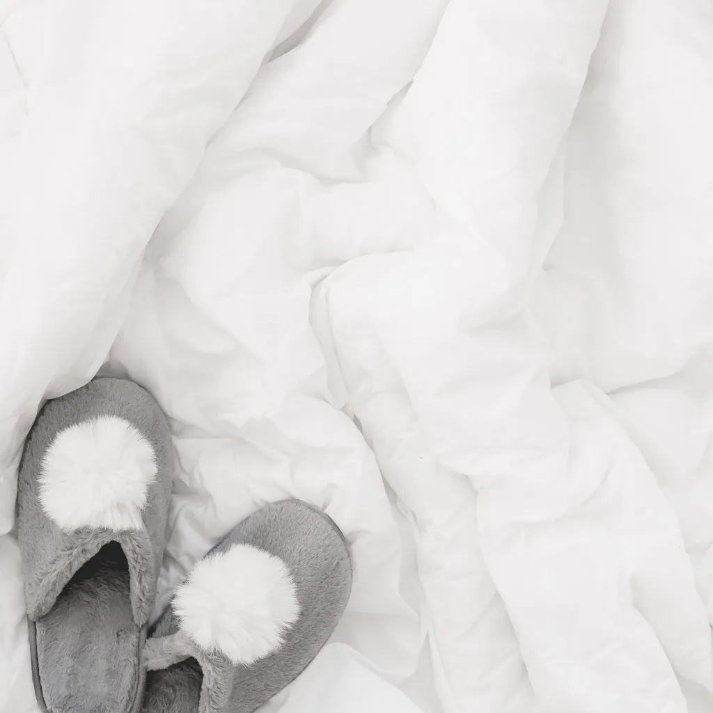 white comforter bunched up with fuzzy grey slippers on bed