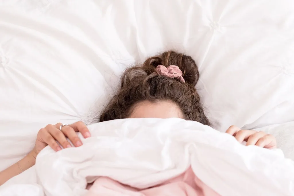 woman hiding under pink blanket and white sheets laying on white pillow
