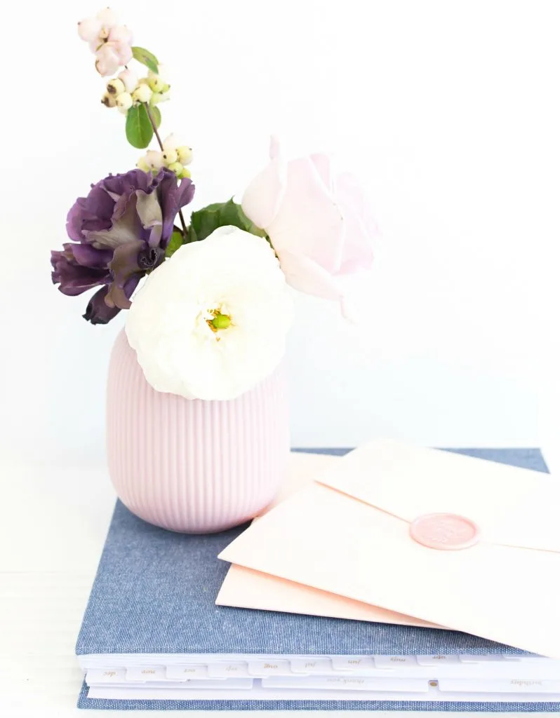 pink envelope on blue journal next to purple vase filled with fresh flowers