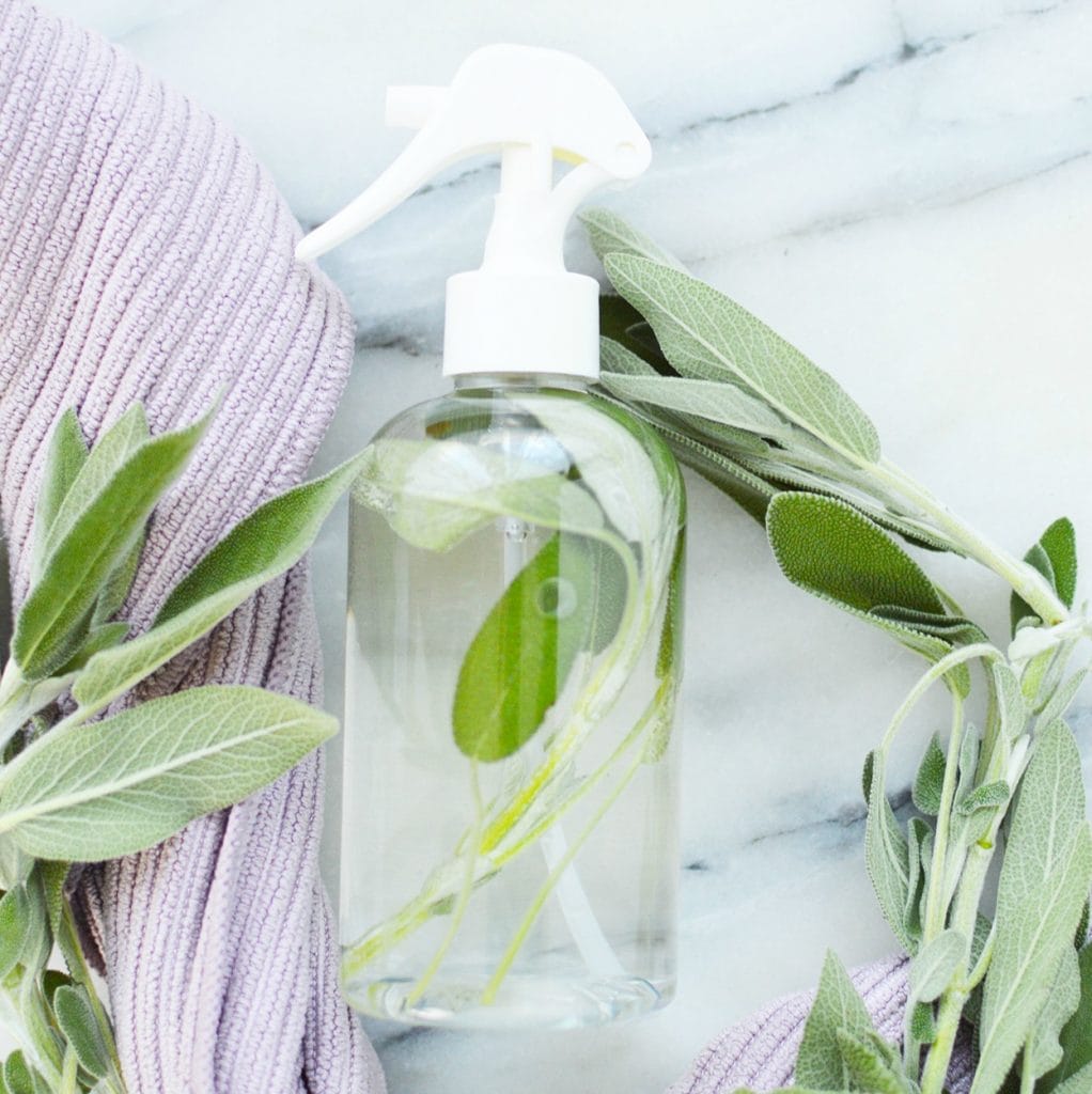 glass clear bottle with diy sage spray real herb leaves against purple cloth and marble background