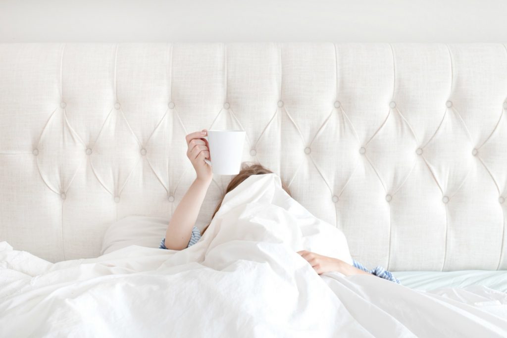 woman hiding under white sheets in bed holding up coffee cup