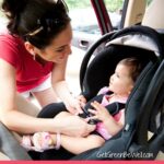 Happy smiling mother placing baby in car seat and closing belt for safety.