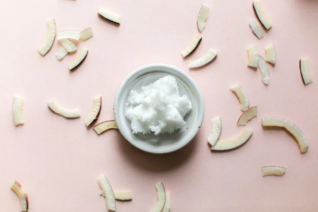 coconut oil in white bowl on pink background surrounded by coconut chips