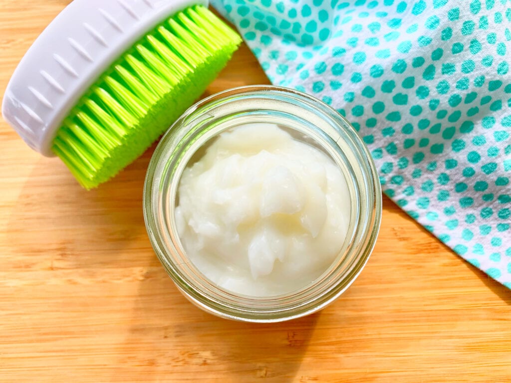 glass jar of gel cleaner on wood table with scrub brush and towel