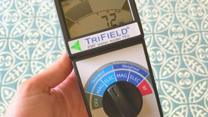 A hand is holding a Trifield EMF meter, model TF2, which is showing a reading on its display. The dial on the EMF meter has various measurement settings for standard, weighted, magnetic, electric, and RF.