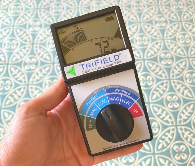 A hand is holding a Trifield EMF meter, model TF2, which is showing a reading on its display. The dial on the EMF meter has various measurement settings for standard, weighted, magnetic, electric, and RF.