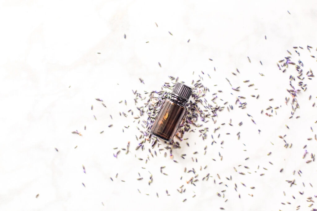 brown glass bottle laying on purple lavender flower buds against white background
