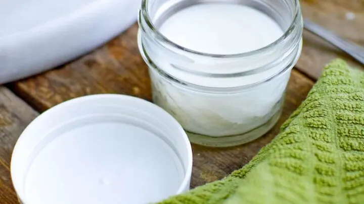 Open glass jar with a white substance inside, next to a white lid and a green textured cloth, all placed on a wooden surface—perfect for showcasing your beeswax furniture polish.
