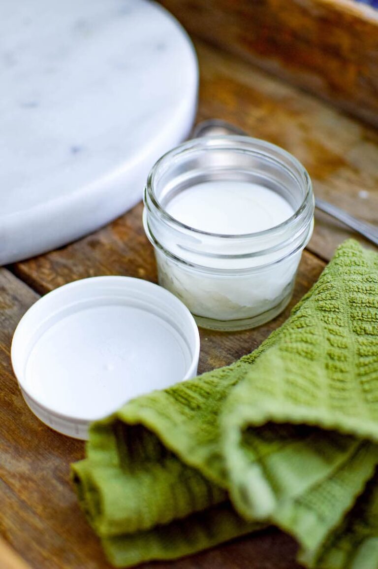 Open glass jar with a white substance inside, next to a white lid and a green textured cloth, all placed on a wooden surface—perfect for showcasing your beeswax furniture polish.