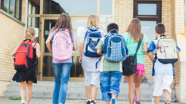 Six children wearing backpacks walk towards a school building entrance, perhaps discussing how to wash a backpack without damaging its fabric.