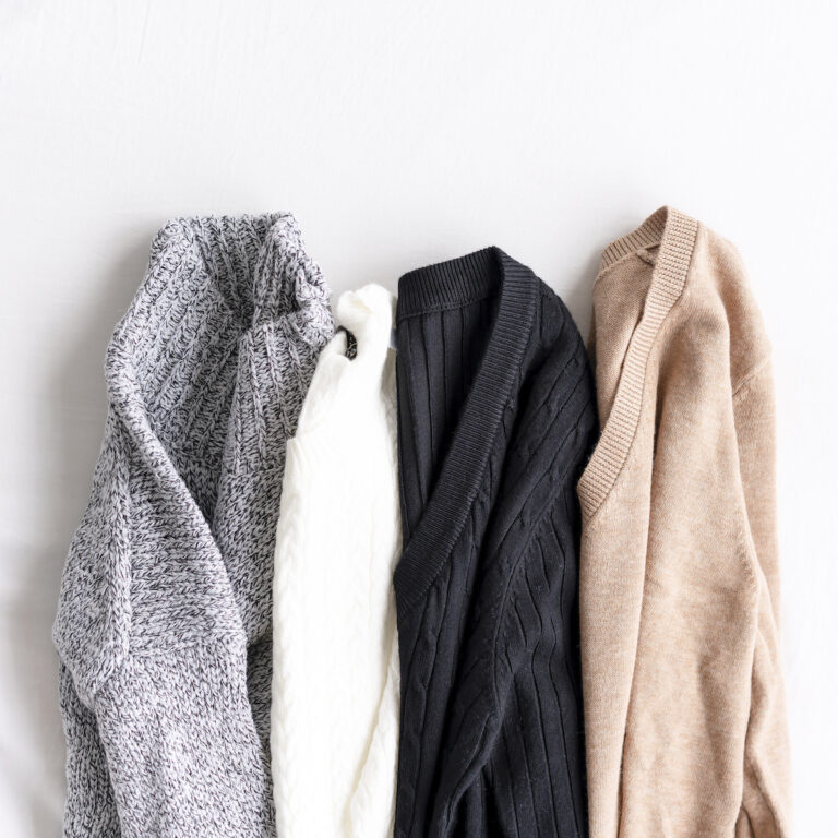 Four knitted sweaters in gray, white, black, and beige are laid out in a row on a white surface. Wondering how to get the smell out? These cozy essentials are as fresh as they look!