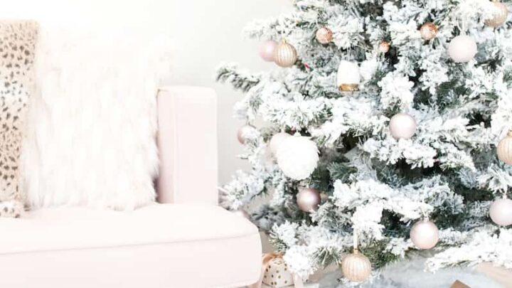 A snowy Christmas tree decorated with pink ornaments stands next to a light-colored sofa with throw pillows, evoking cozy holiday vibes. However, be cautious as certain allergens from the tree could potentially make you sick. Wrapped gifts are placed lovingly under the tree.