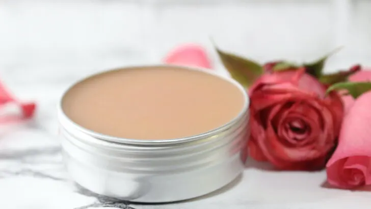 small aluminum tin filled with pink colored rose salve on marble background with fresh rose laying beside it