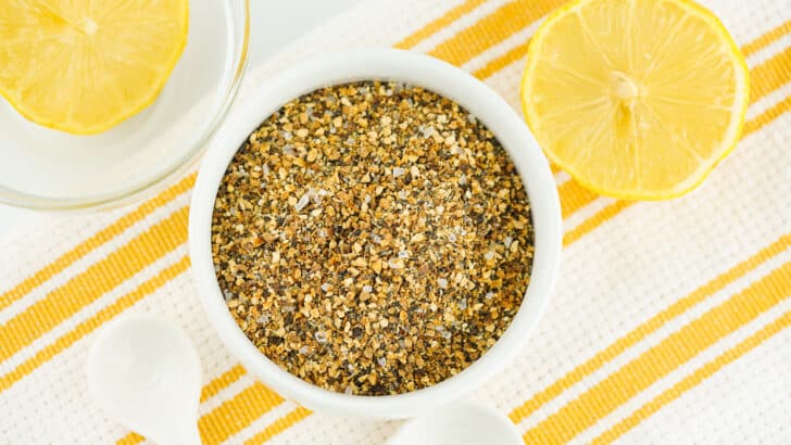 A bowl of homemade lemon pepper seasoning with spoons and lemons next to it.