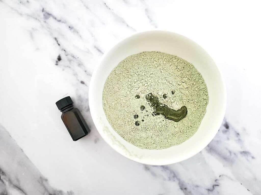 A bowl of green clay with a bottle of essential oil next to it to make Detox Clay Bath.