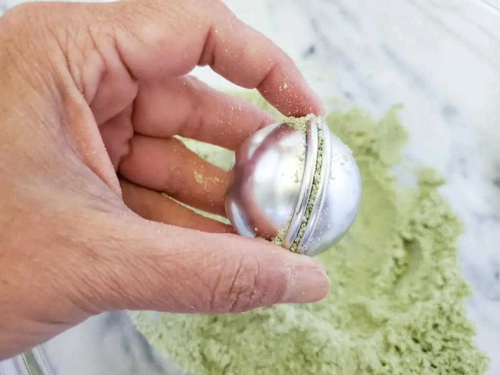A hand is holding a green lemongrass bath bombs powder in a bowl.