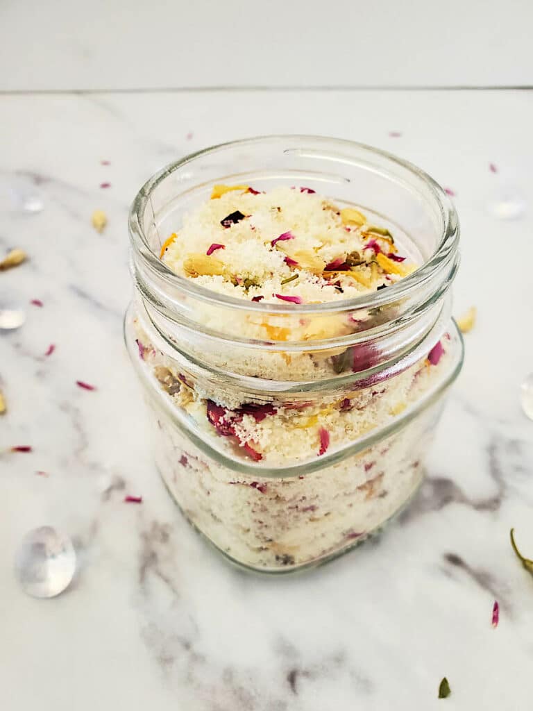 A jar of milk and honey bath soak ingredients in a glass jar on a white table.