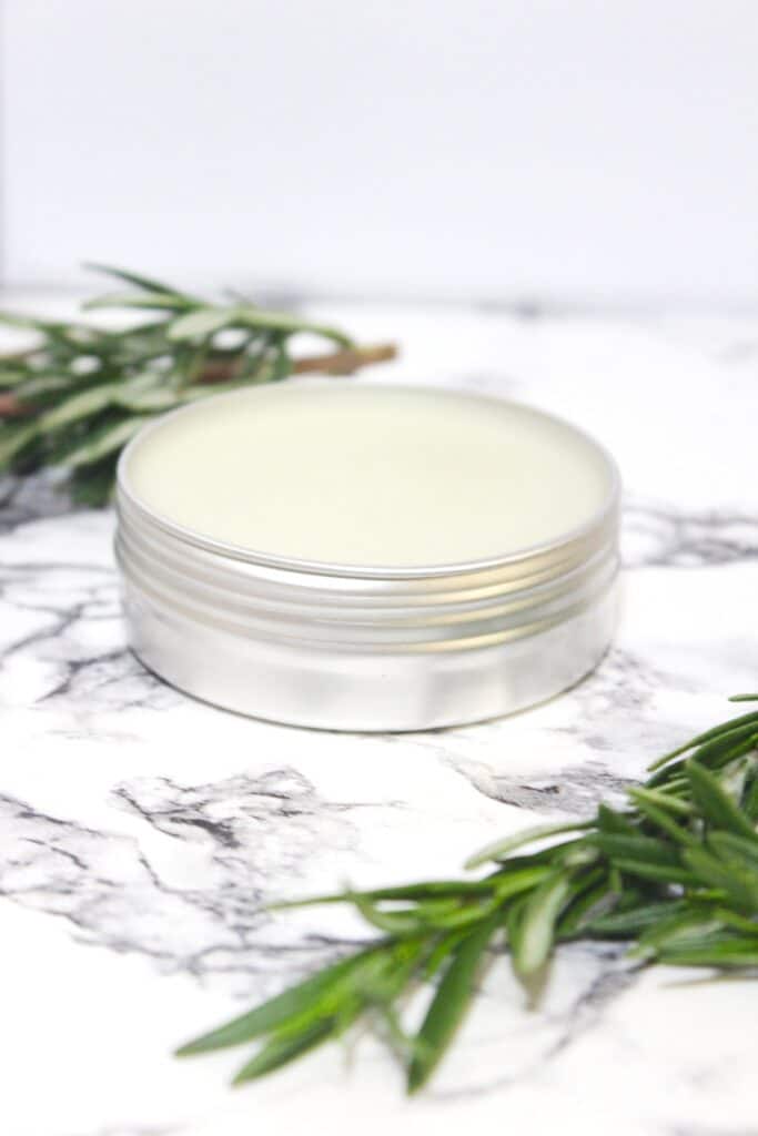 A tin of natural Rosemary Salve with rosemary sprigs on a marble table top.