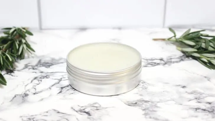 A metal tin of Rosemary Salve with rosemary sprigs on a marble table.