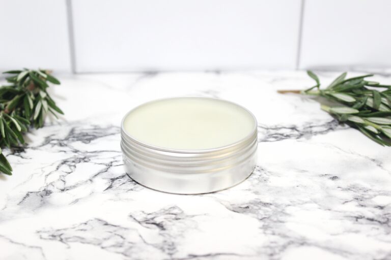 A metal tin of Rosemary Salve with rosemary sprigs on a marble table.