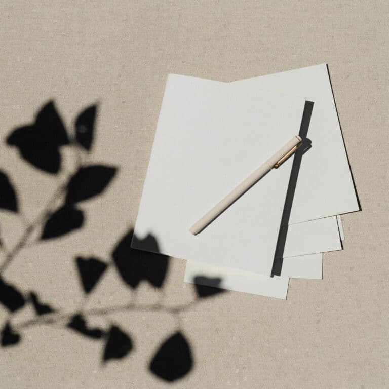 A pen rests on a stack of blank white paper, providing a sense of focus for beginner tips for minimalism as the shadow of a plant is cast across the surface.