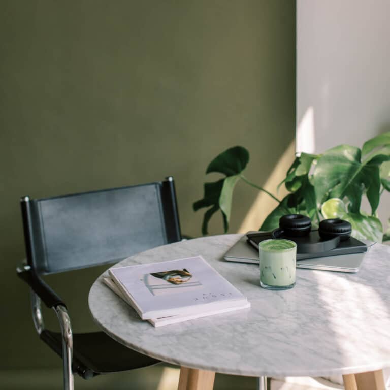 An iced matcha latte, a magazine, and a laptop with headphones sit on a round marble table next to a chair, reflecting a setting of minimalist habits.