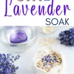 A bowl filled with a mixture of oats and lavender flowers. Dried lavender flowers are scattered nearby, and a glass bowl with a purple candle is in the background. Text reads, "Homemade Oat Bath Soak.